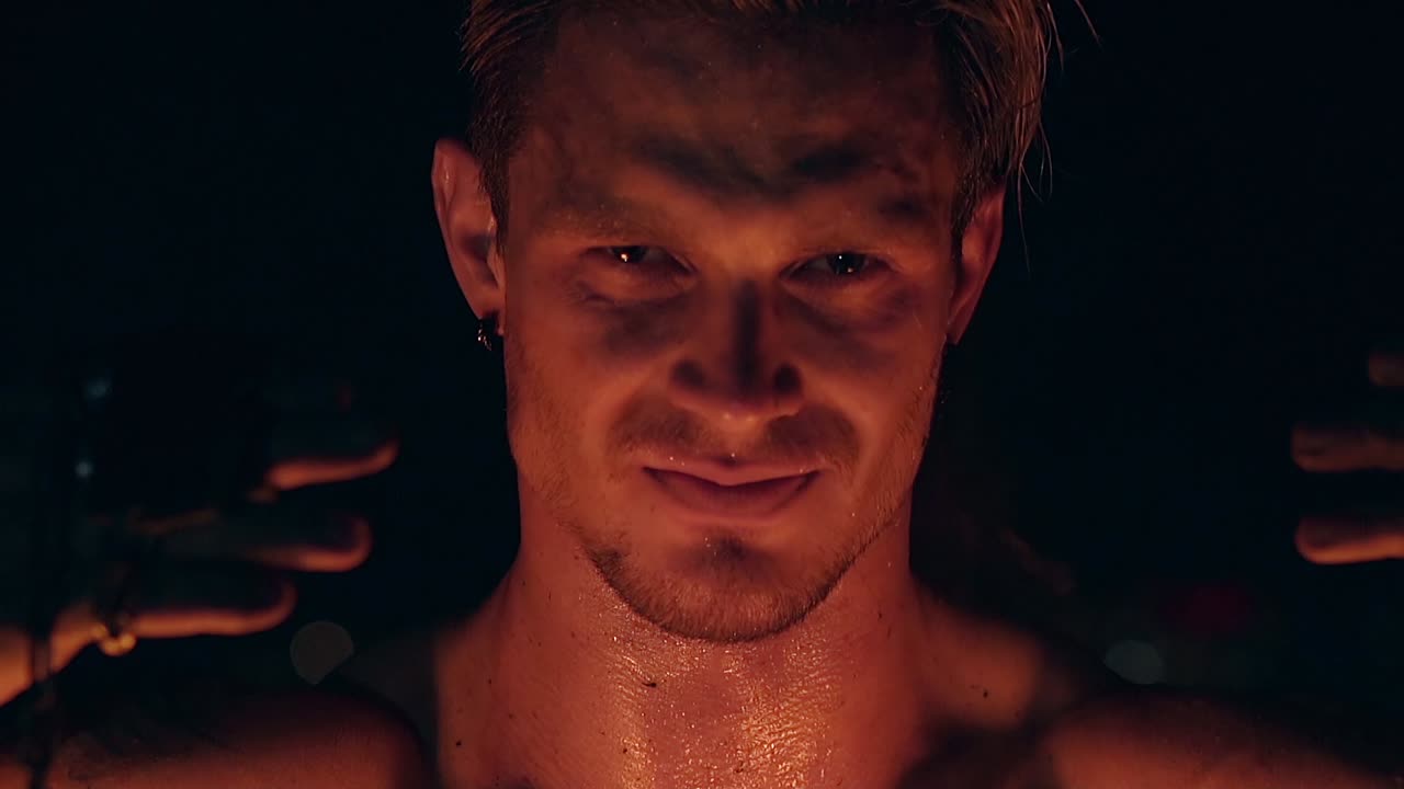 젊은 금발 남자가 두 마리의 포를  ⁇ 사슬에 매달고 남자가 카메라를 쳐다보며 피부에 불을 반사합니다.