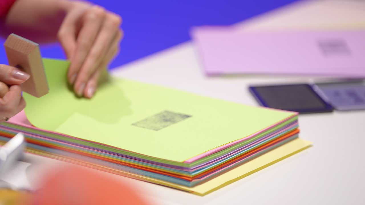 A female hand presses a wooden stamp onto a vibrant stack of pastel papers in a colorful retro office. The scene highlights a detailed and tactile moment in a stylish workspace with soft lighting