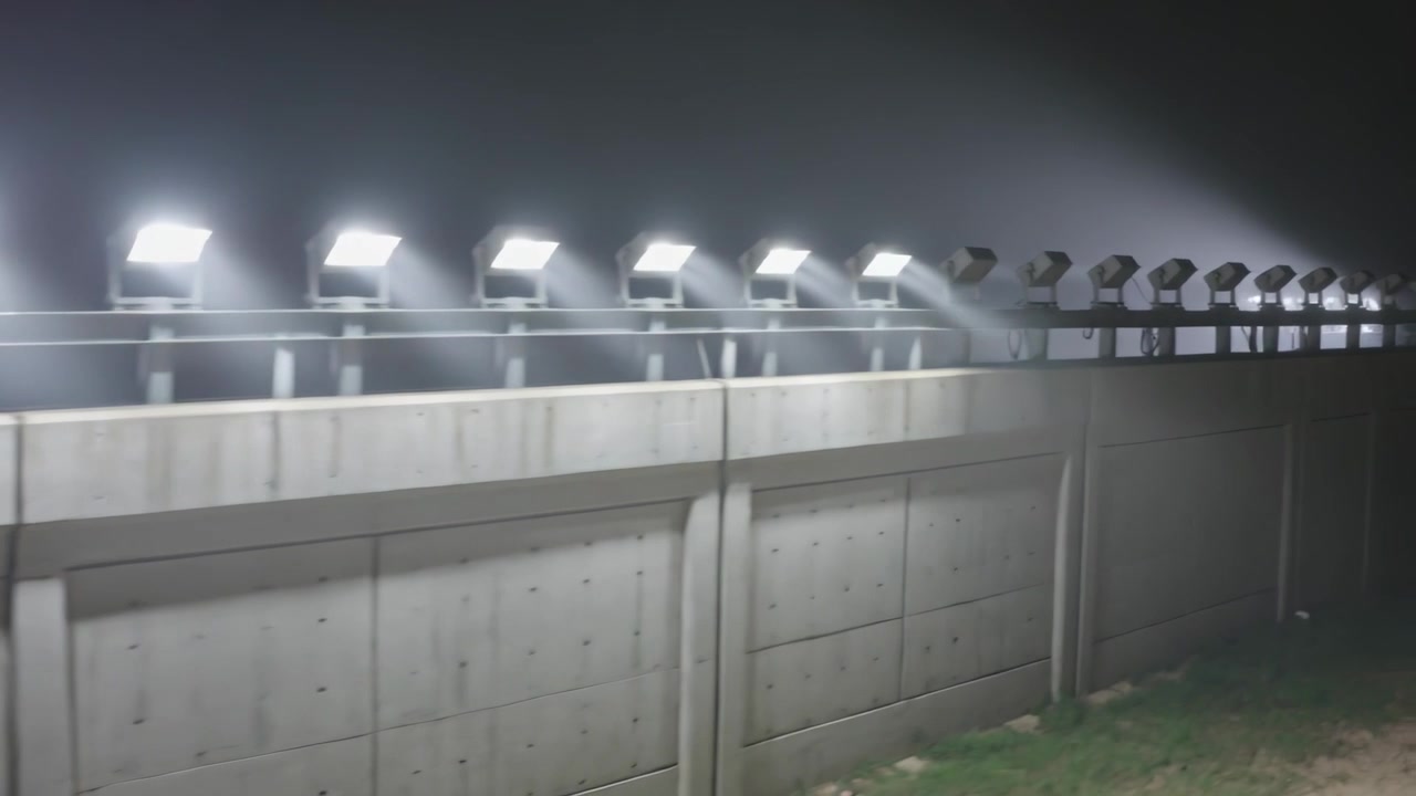 Siklus daya deretan lampu sorot di malam hari, membentuk berkas cahaya yang terlihat untuk keamanan perimeter.