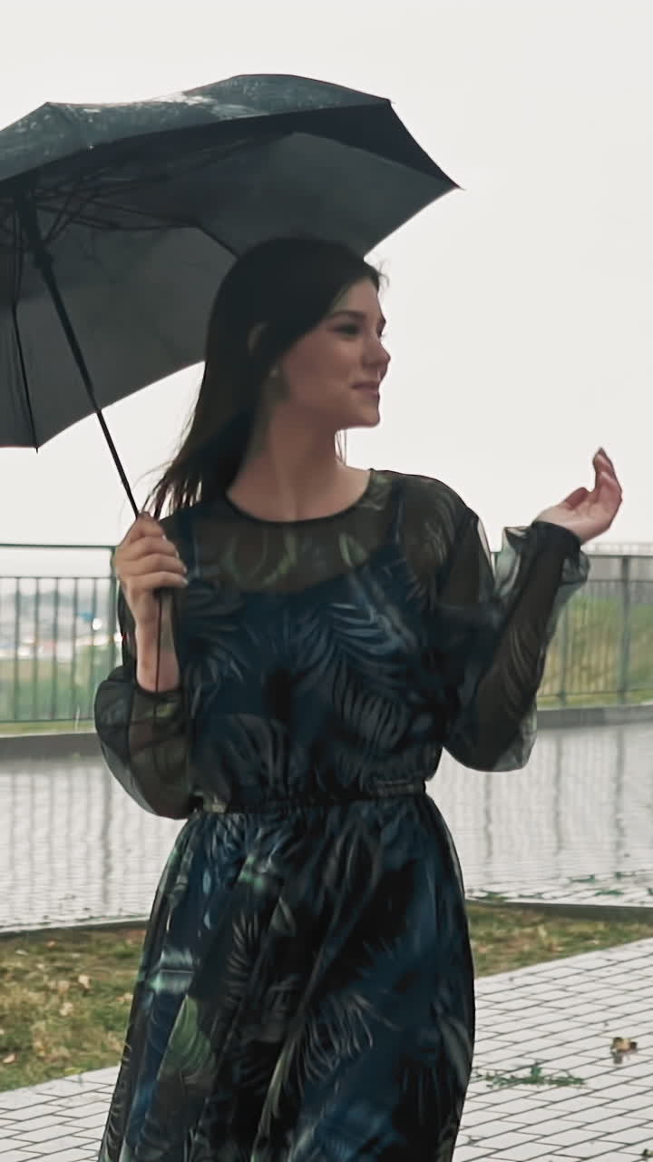 Attractive cheerful lady in elegant dress holds black umbrella catching falling rain drops by palm on city park viewpoint on spring day slow motion
