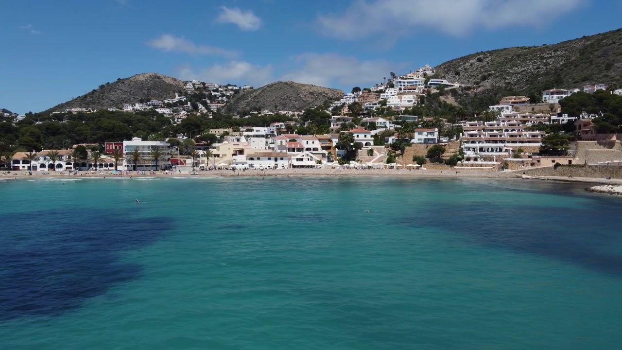 Orbita mozzafiato di un drone che mostra le acque turchesi incontaminate, la baia panoramica e le sabbie dorate della spiaggia di El Portet a Moraira, Spagna, catturata da una prospettiva aerea elevata.