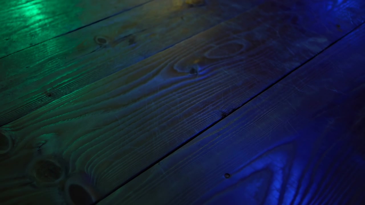 Low oblique close-up of diagonal wooden planks showing grain, knots, seams, nail heads, scratches, and worn finish while directional colored light creates green, teal, blue gradients