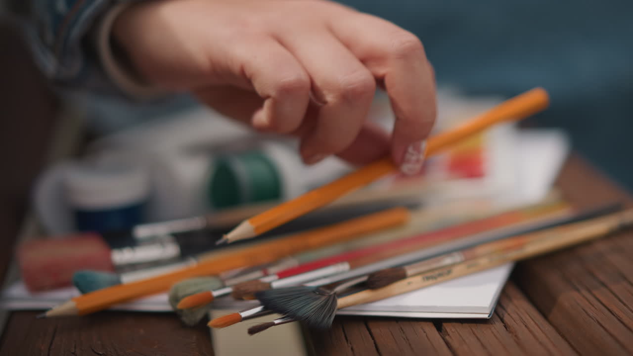 Closeup art supplies on wooden table, artist hand reaching for brush amid scattered pencils and paint jars, palette with vivid swatches, shallow depth of field, warm natural light, relaxed creative