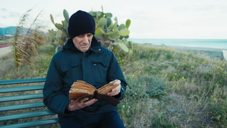 elderly man reading a novel on a bench near the sea