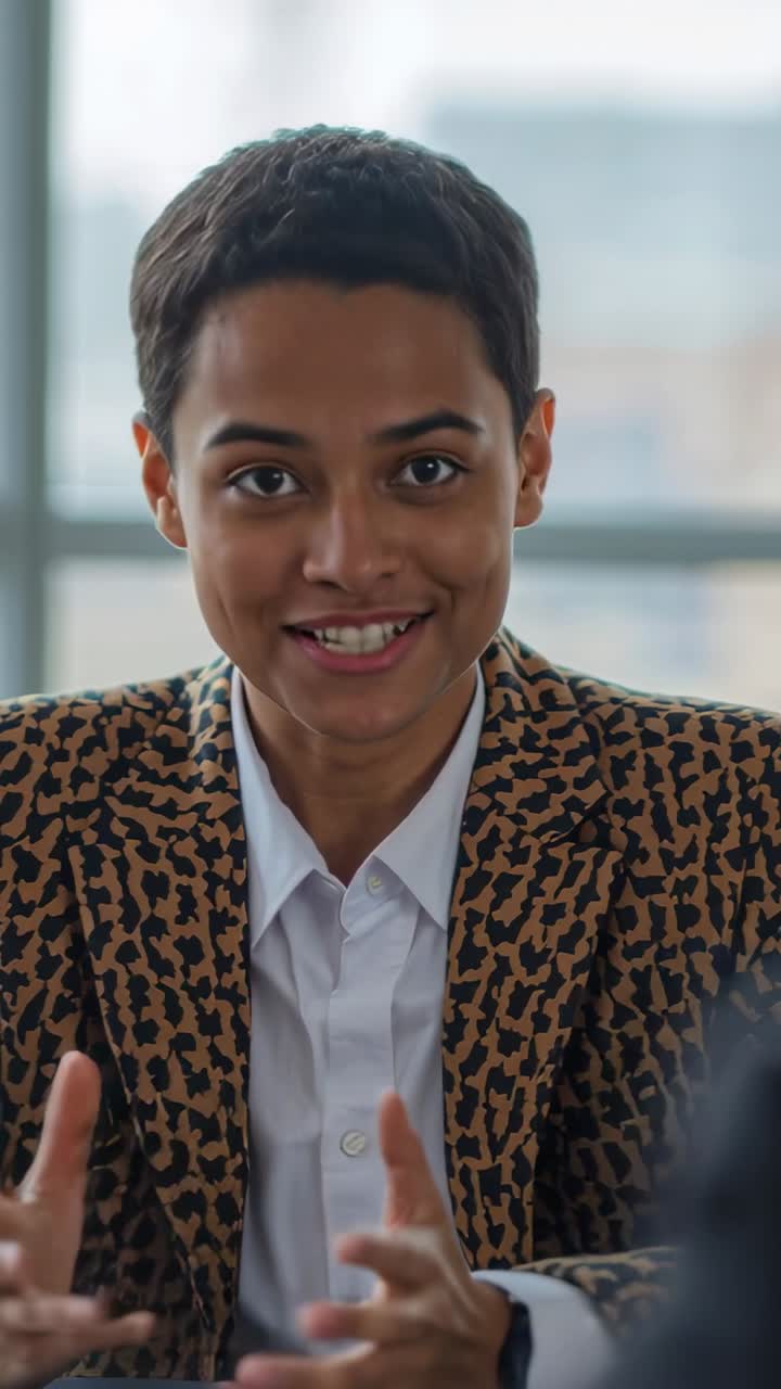 Video verticale: Donna che sorride a un segnale fuori campo, parla a un tavolo in ufficio indossando un blazer leopardato.