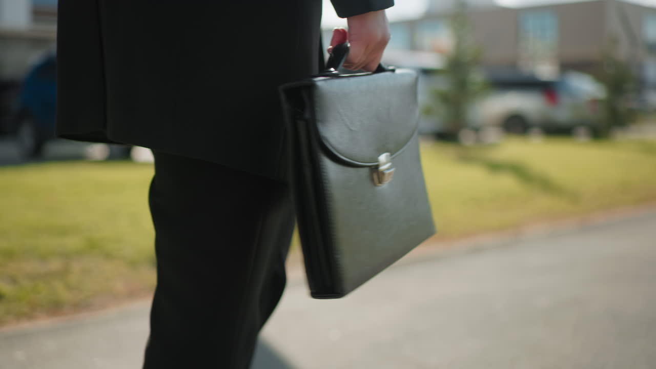 Back view of individual in black coat walking outdoors, holding briefcase firmly, surrounded by blurred cars, greenery, and modern glass buildings in urban setting under bright sunlight