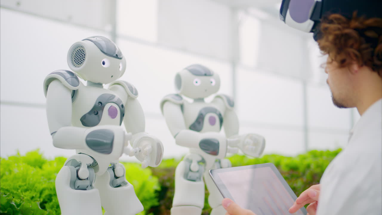 Laboratory technician in a white coat wearing virtual reality headset analysing graphs on a tablet while interacting with two humanoid robots in a greenhouse farm