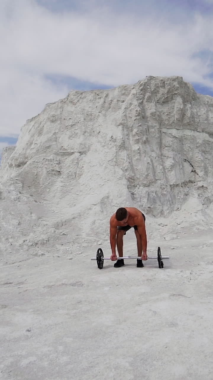 Strong shirtless athlete taking heavy barbell outdoors. Muscular man lifting barbell on white mountain background. Slow motion. Vertical video