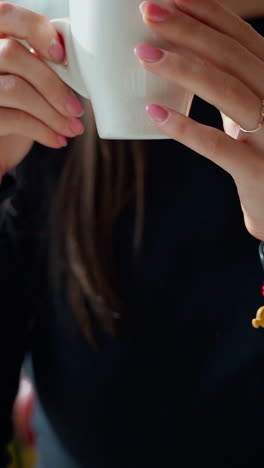 Beautiful girl drinking coffee in a cafe. Young woman sitting at the table near the window and enjoys hot drink in the restaurant. Slow motion. Vertical video