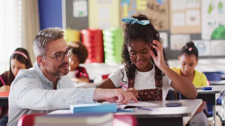 Diverse happy male teacher helping schoolgirl sitting in classroom during learning