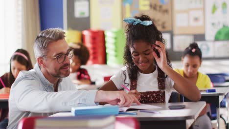 diverse happy male teacher helping schoolgirl sitting in classroom during learning