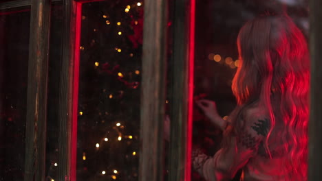 Long-haired woman decorating Christmas tree in the beautiful village house. Decorated room with red lights and preparation for the Christmas holidays.