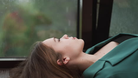 a young woman in a green jacket lies down by a window, looking away with a pensive expression. rain falls outside.