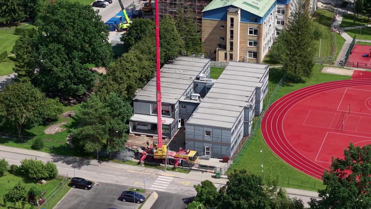 Luftoptagelser viser en kran, der samler modulbygninger ved siden af en levende rød sportsbane på en solrig dag, omgivet af træer, byarkitektur og frodigt grønt