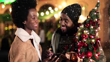 Vista-De-Cerca-De-Una-Alegre-Pareja-Afroamericana-Hablando-Y-Viendo-Algo-Por-Teléfono-Mientras-Nieva-En-La-Calle-En-Navidad