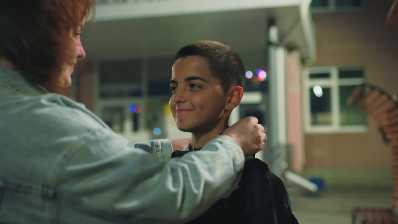 Mother holds boy’s backpack straps while speaking to him outside school entrance during early morning, expressing care, encouragement, and support before class begins under soft exterior lighting