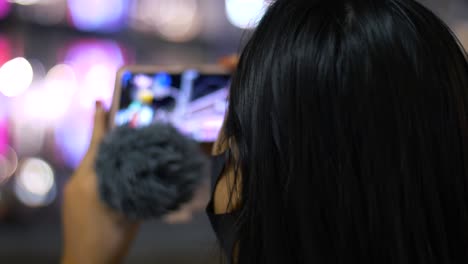 slow motion close up head shot with bokeh of a young, asian female tourist wearing a face mask, vlogging with her smartphone and speaking into a mic in a city at night