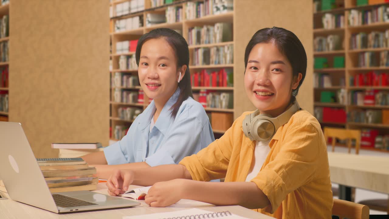 Asian Woman Students With Headphones Smiling And Showing Thumbs Up Gesture To The Camera While Reading Books On The Table With Laptop In The Library
