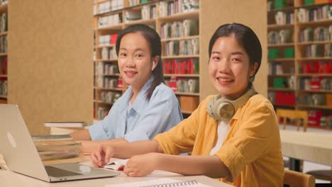 asian woman students with headphones smiling and showing thumbs up gesture to the camera while reading books on the table with laptop in the library