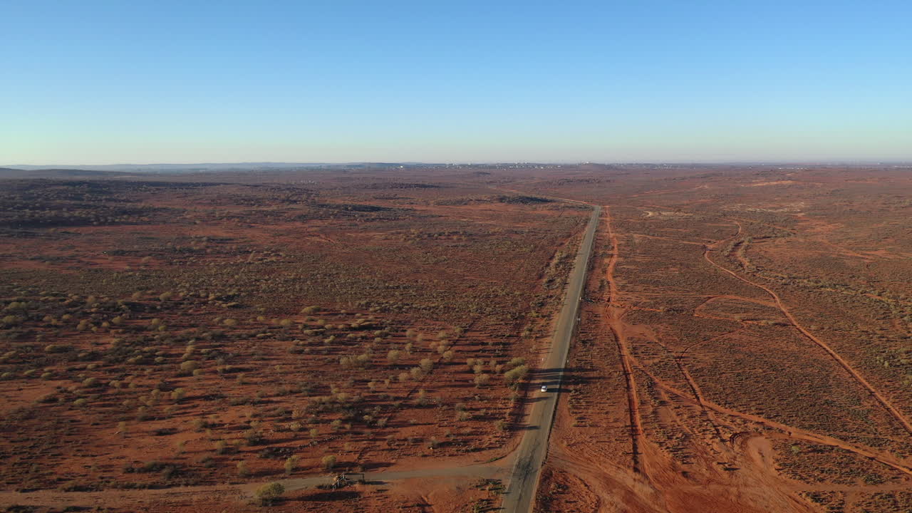 항공:오스트레일리아 부러진 언덕 근처의 붉은 흙 아웃백으로 둘러싸인 활주로를 따라 운전할 때 멀리 있는 흰색 차량을 추적하는 높은 각도의 드론 샷