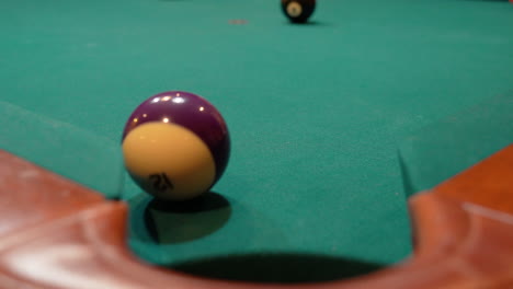 pool balls are broken on an 8 ball triangle rack and the purple stripe 12 ball slowly rolls towards edge of corner pocket, closeup with solids and stripes scattering across the table with green felt