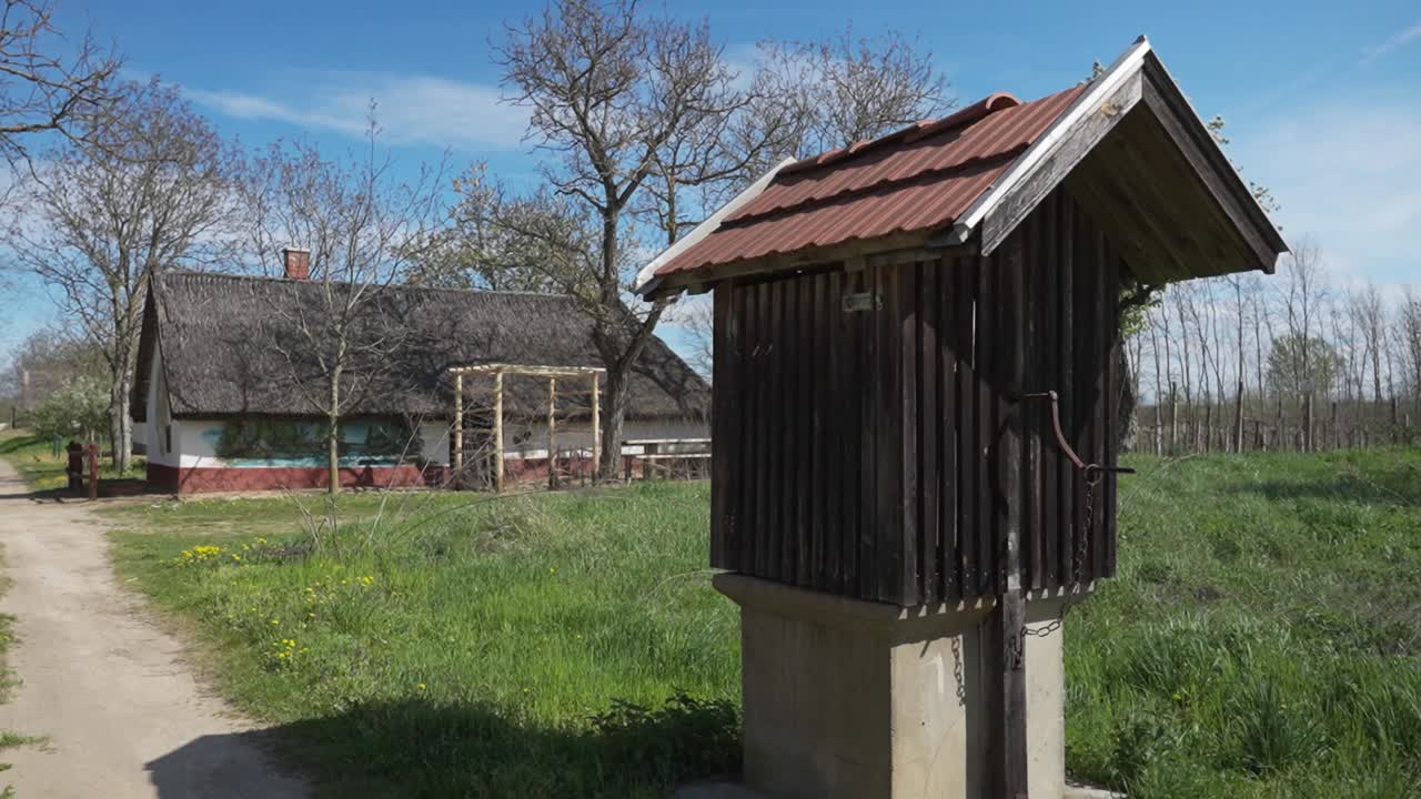 Traditional wooden water well in green field with thatched house nearby