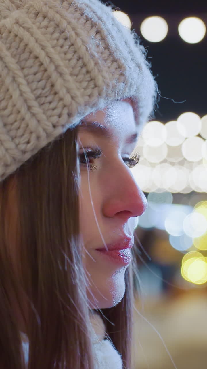 Young woman wearing beanie and warm coat brings hands close to mouth, exhaling to warm them on a cold winter night, city lights, festive atmosphere, and people in background