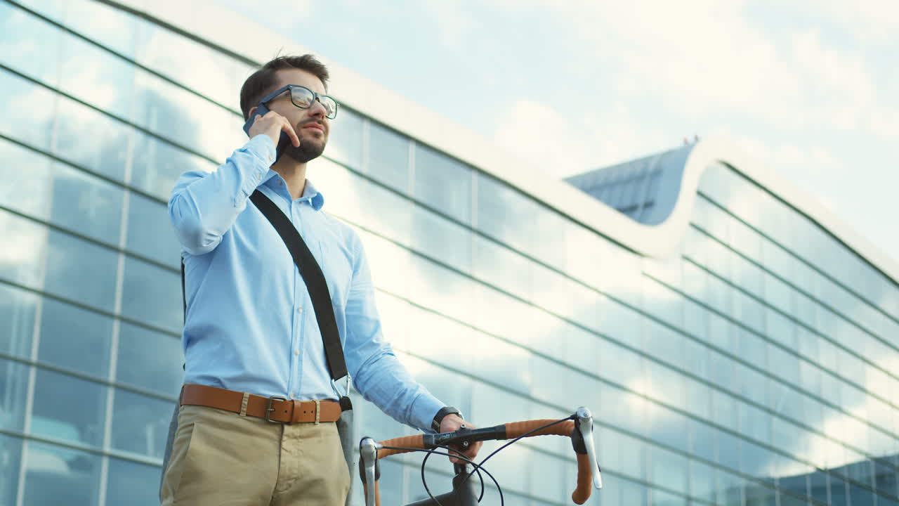 стильный мужчина стоит рядом с велосипедом, разговаривает по мобильному телефону и улыбается возле большого здания