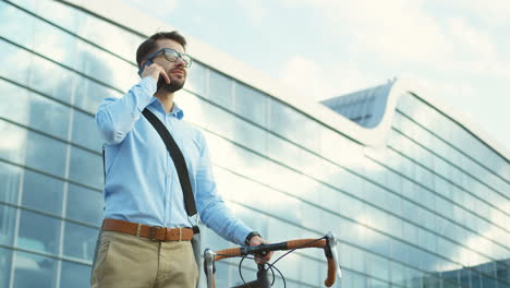 Stylish-man-standing-close-to-his-bicycle,-talking-on-the-mobile-phone-and-smiling-outside-a-big-building