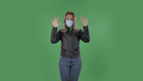 의료 보호 얼굴 마스크를 입은 아름다운 젊은 여성의 초상화는 카메라를보고 주먹을 보여 주고, 한, 두, 세, 네, 다섯 손가락을 세고, 검은색 재과 청바지를 입은 느슨한 머리카락을 가진 금발은 스튜디오에서 녹색 화면에 건강 보호.