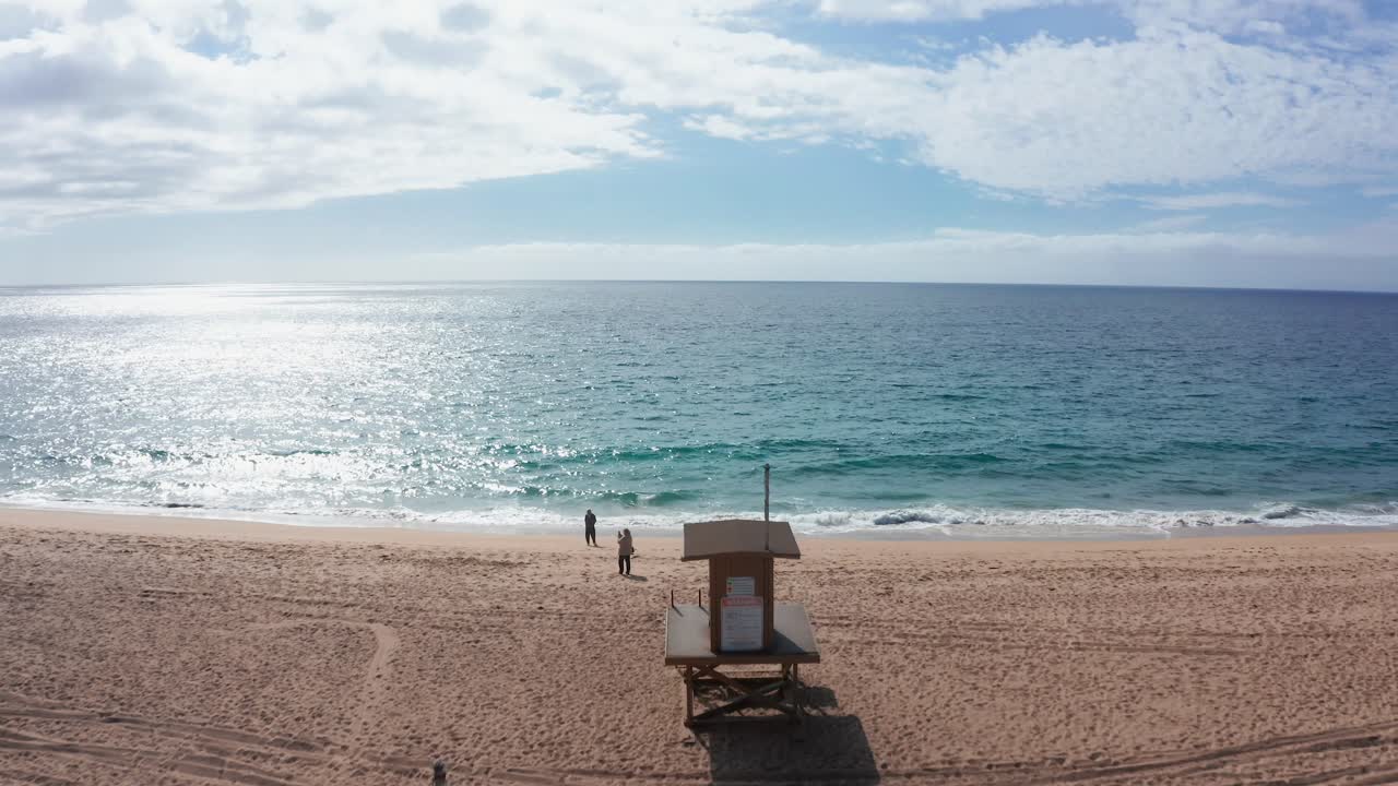 vuelo de bajo nivel sobre la playa de arena con cabina de salvavidas y mar azul, iluminado por la luz del sol