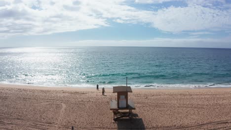 low level flight over sand beach with lifeguard cabin and blue sea, lit by sunlight