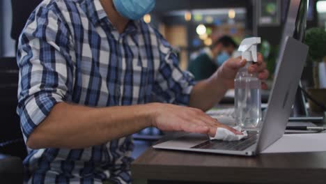 Caucasian-man-wearing-face-mask-cleaning-his-laptop-using-tissue-and-disinfectant-spray-at-modern-of