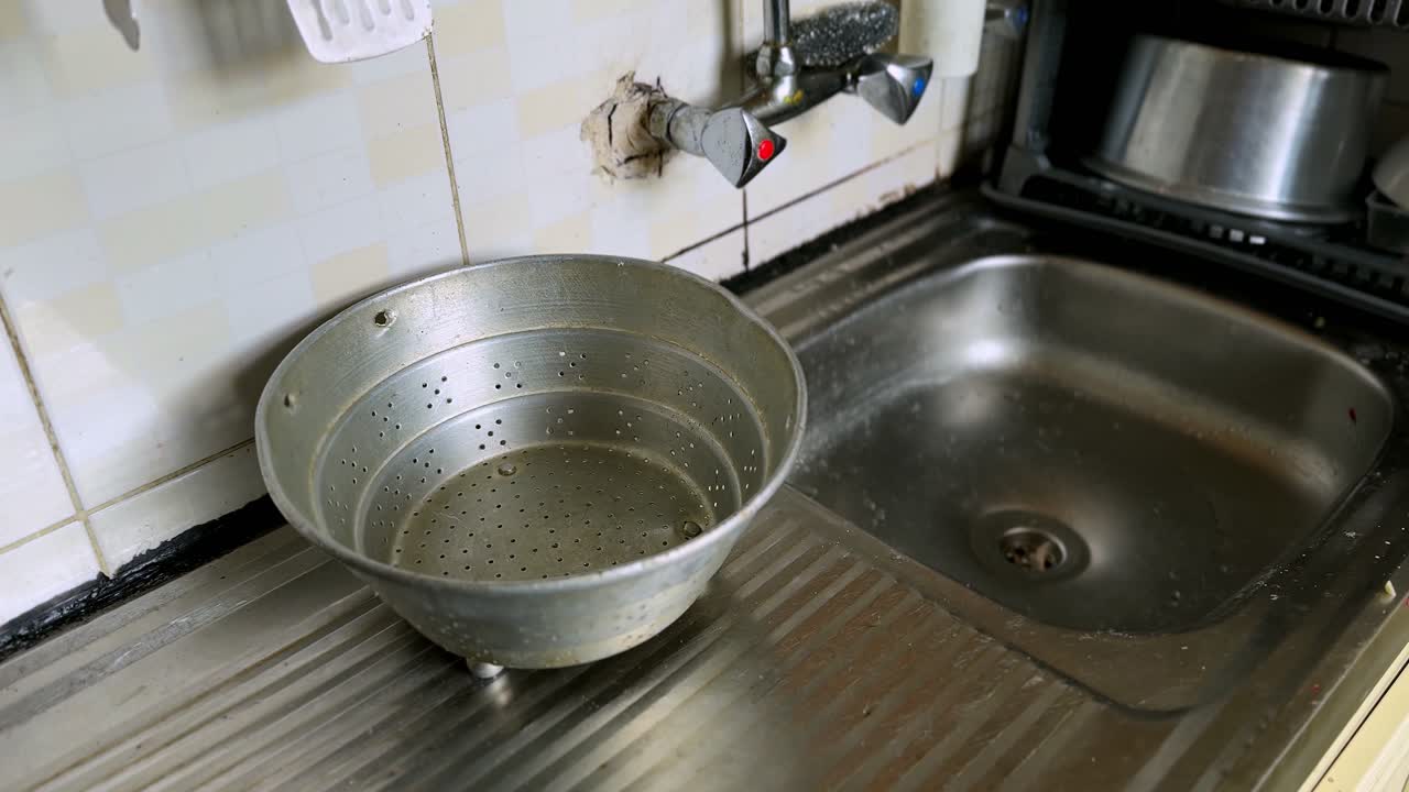 Gammel perforeret metalsi placeret på et drænfelt af rustfrit stål ved siden af en køkkenvask med vægmonteret armatur og plettede flisevægge i naturligt indendørs lys.
