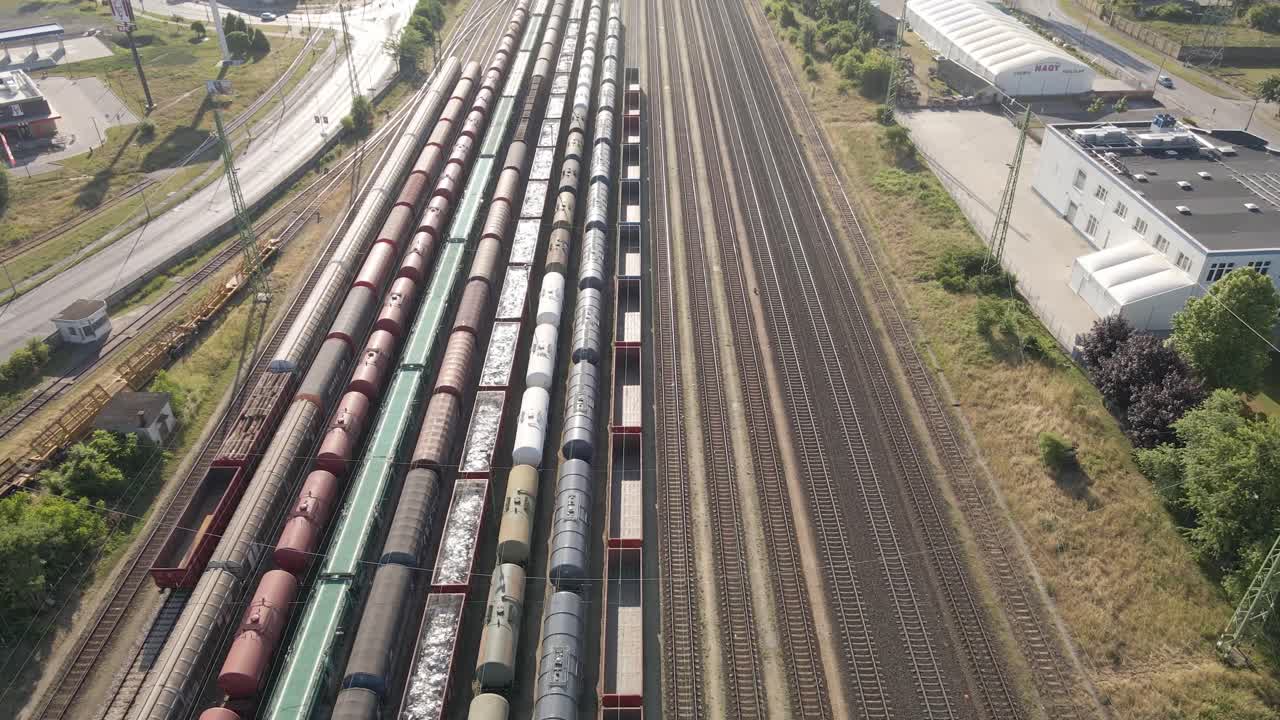 Aerial view of a railway station with multiple freight trains parked on the tracks. The surrounding area includes industrial buildings, roads, and warehouses, showcasing a logistics hub.