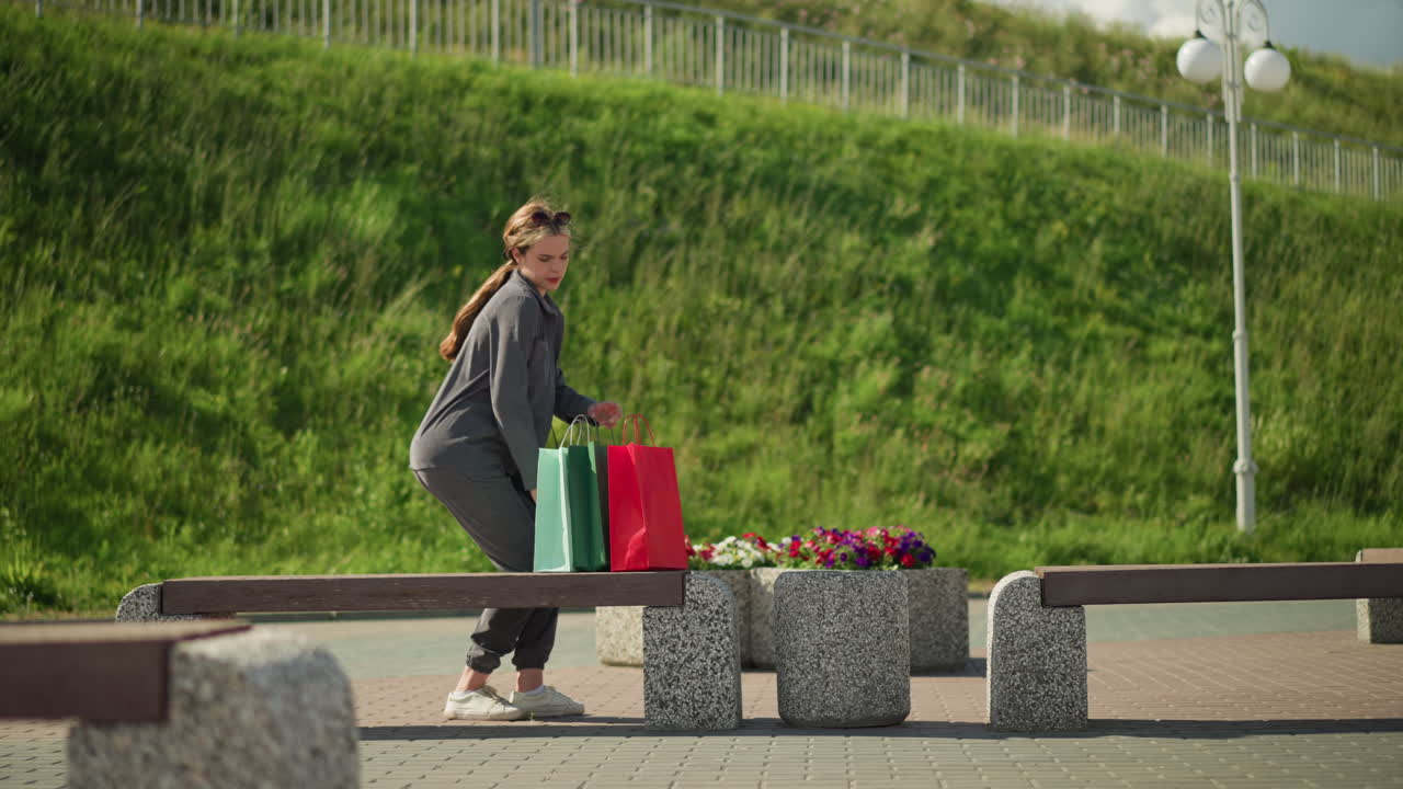 Lady places colorful shopping bags on a bench, sits down, and relaxes under sunlight reflecting on her face, blurred background features a street lamp and greenery