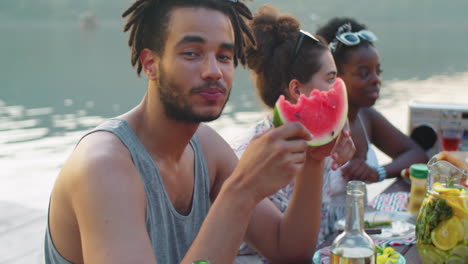 young man eating watermelon and smiling at camera at lake party