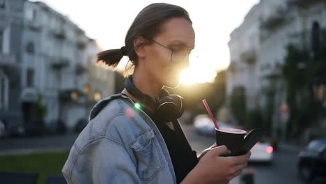 pretty young girl in blue shirt and headphones on her neck standing on the city street and scrolling her mobile phone. holding a