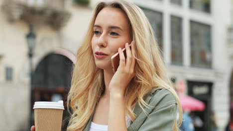 vista de cerca de una mujer rubia caucásica hablando por teléfono inteligente mientras camina por la calle y bebe café para llevar