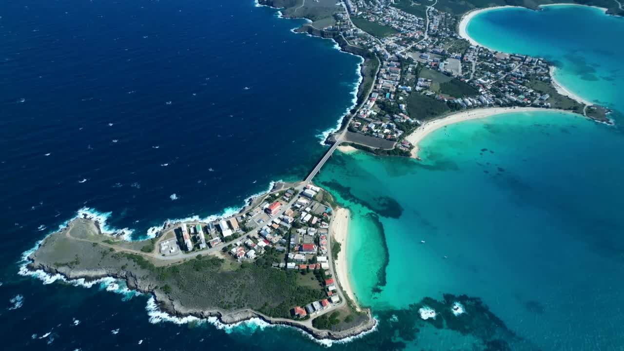 Drone in decollo, che sorvola una penisola in una baia costiera, mostrando una strada rialzata, tetti rossi e il contrasto tra oceano e baia.