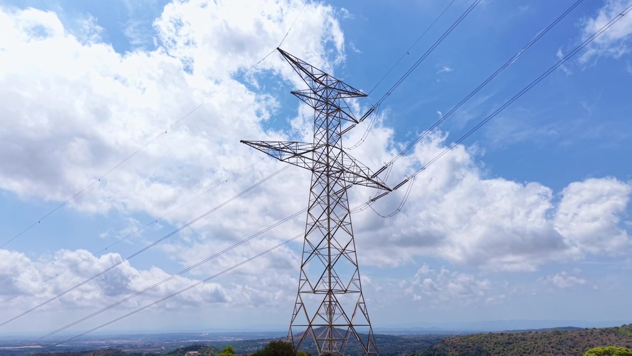 Tall steel electricity transmission tower rising against blue sky with white clouds, power lines stretching across rural landscape with green hills in background