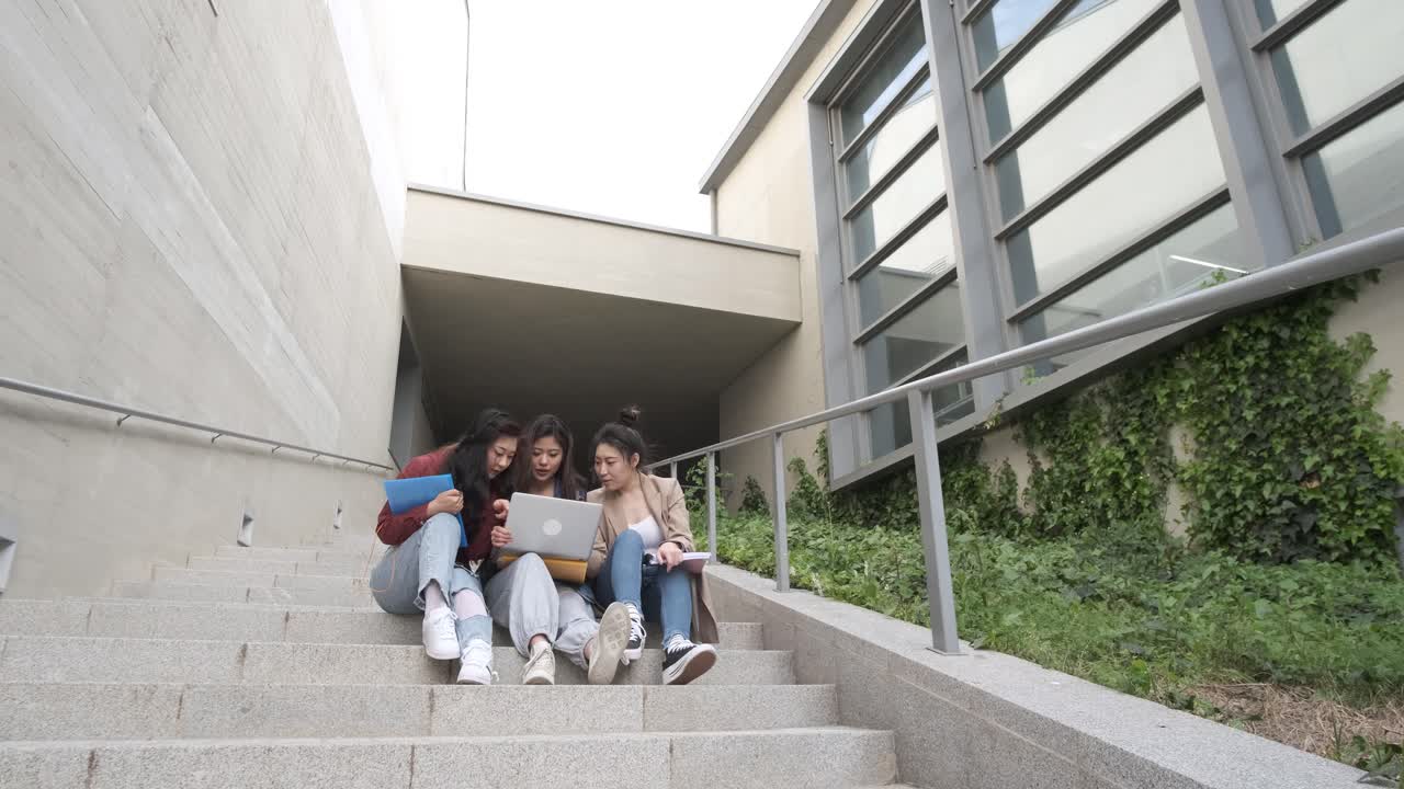 Group of Asian female students using a laptop and studying together while sitting on the stairs of the university campus.