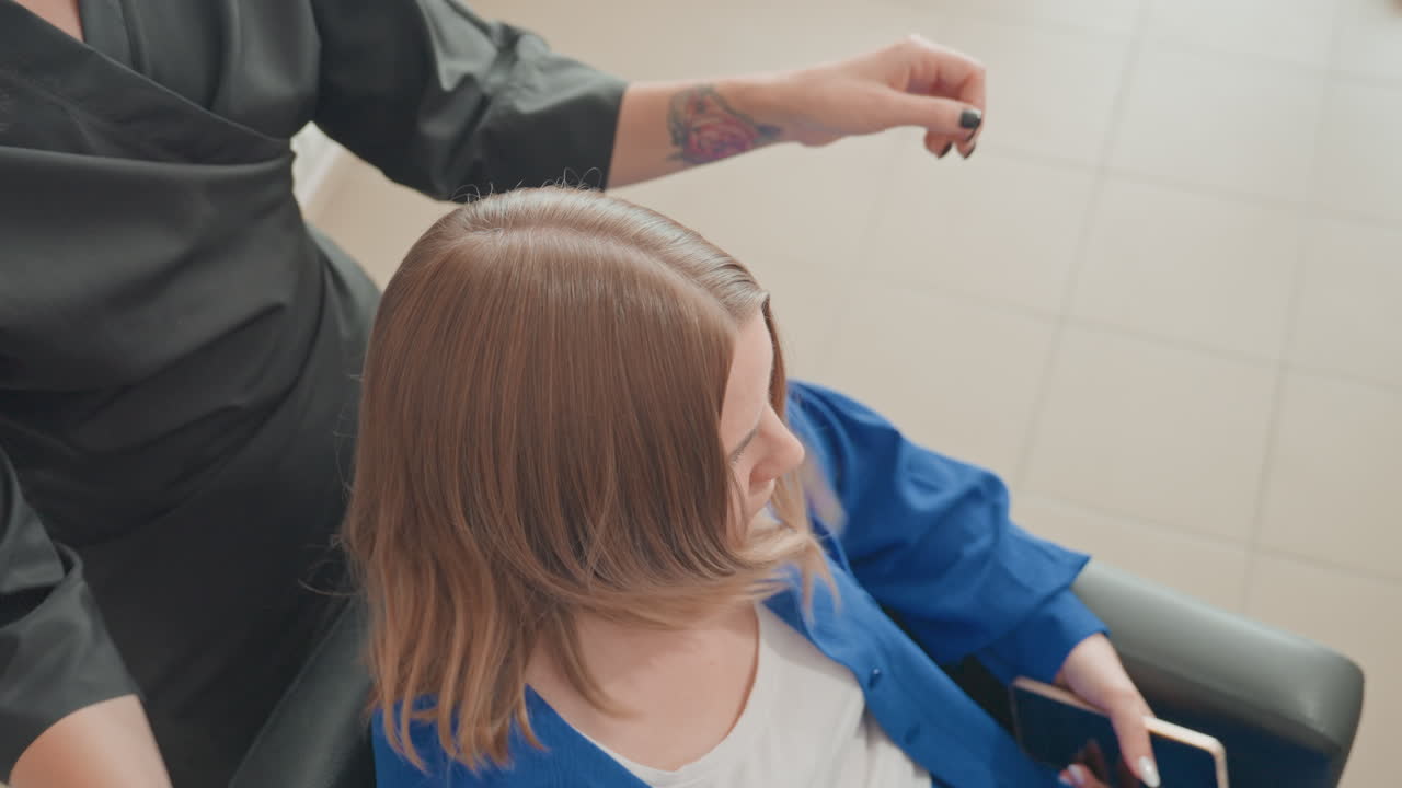 Primer plano cenital de una mujer caucásica sentada en el sillón de la peluquería, peinando su cabello castaño claro hasta los hombros con una trenza. El peluquero, en uniforme negro, separa los mechones con las manos. La clienta sostiene un smartphone. Suelo moderno de baldosas.