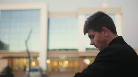 man in black clothing walking out checks time thoughtfully with glass building in background featuring soft light glow, stylish, confident individual in urban environment