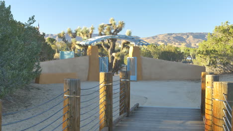 Tilt-up-along-an-empty-wooden-walking-bridge-to-a-blue-metal-gate-in-a-desert-habitat-nature-preserve-with-Joshua-Trees-during-sunrise-golden-hour-in-Antelope-Valley,-California