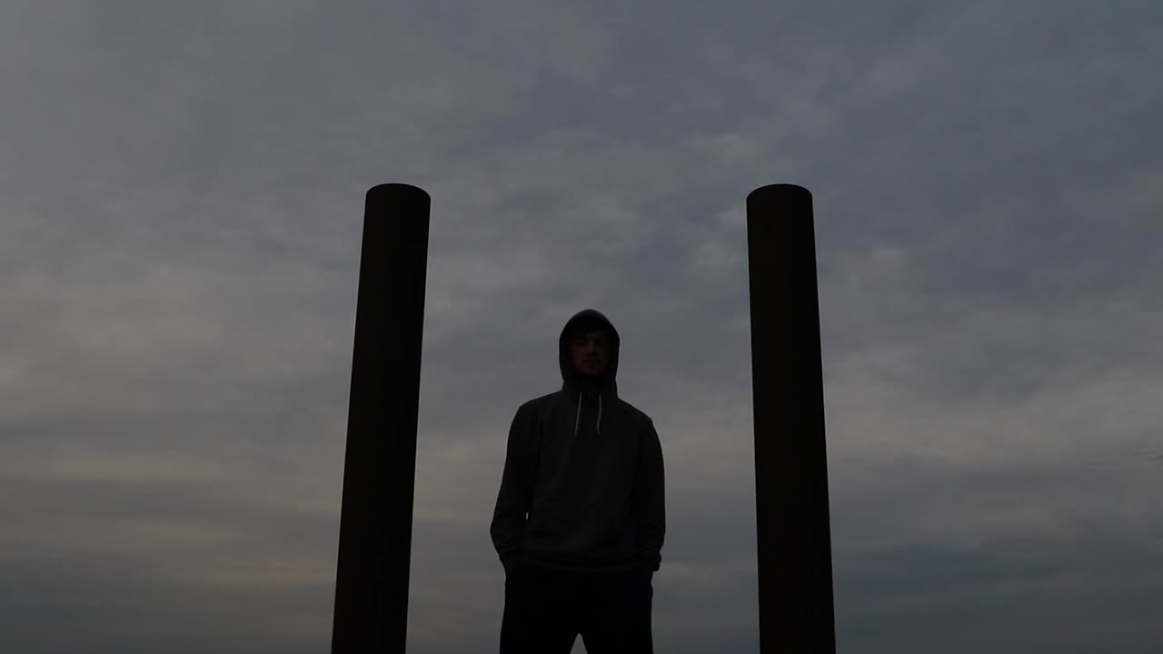 Young caucasian man wearing hoodie stands between two pillars while staring into the distance. Push in slider movement