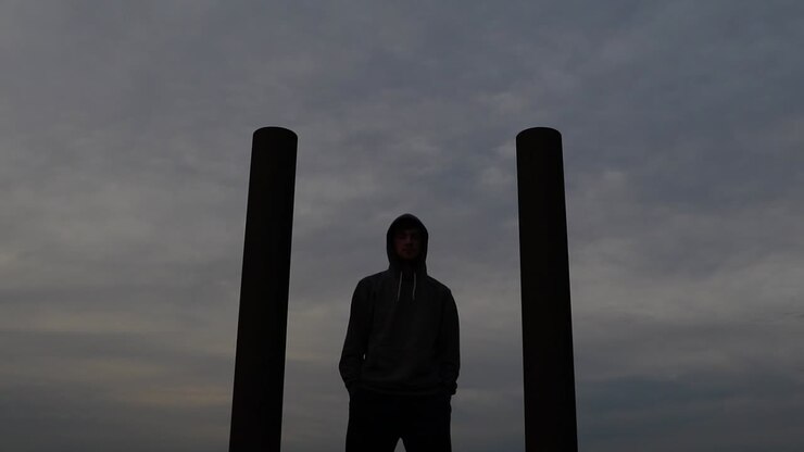 Young caucasian man wearing hoodie stands between two pillars while staring into the distance. Push in slider movement
