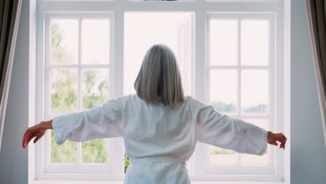 senior woman opens curtains and stretches in front of window