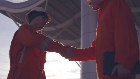 close up view of two construction workers in orange uniform and hardhats shaking hands at the building object. slow motion