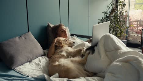 a blonde girl lies on the bed with her dog and watches a movie on a laptop in the morning. holidays with pets